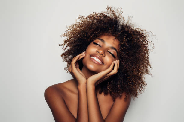 Young black woman with curly natural hair smiling in delight while holding her face. Natural skin, no makeup makeup, natural 4c hair.